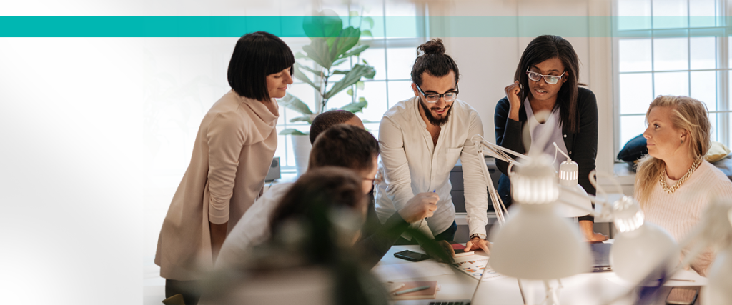 Team of diverse professionals collaborating around a desk in a bright office setting with natural light and greenery.
