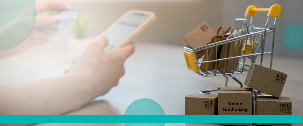 Close-up of a hand using a smartphone for online shopping, next to a miniature shopping cart filled with small cardboard boxes labeled 'Online Fundraising.