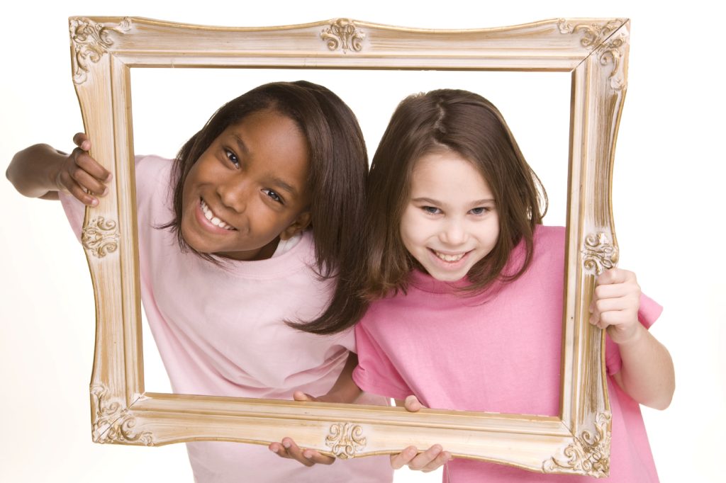 Two smiling girls holding a decorative wooden picture frame, symbolizing friendship and creativity.