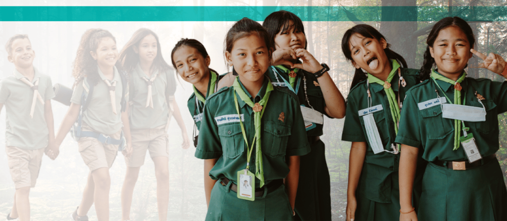 roup of young scouts in green uniforms smiling and posing outdoors, with a forest background and other scouts walking in the distance.