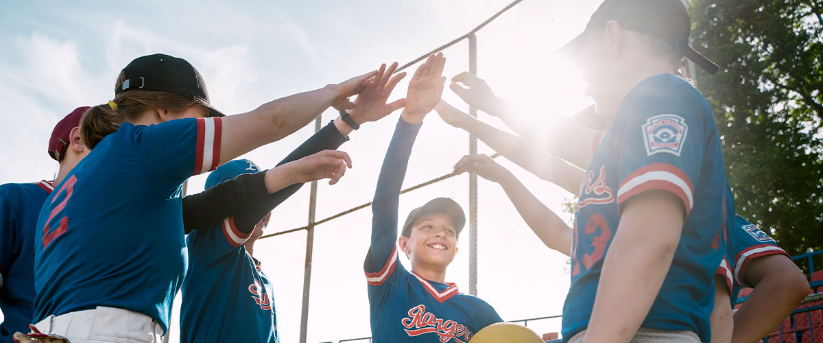 Baseball team in uniforms doing a group cheer with hands together under a bright sunny sky.