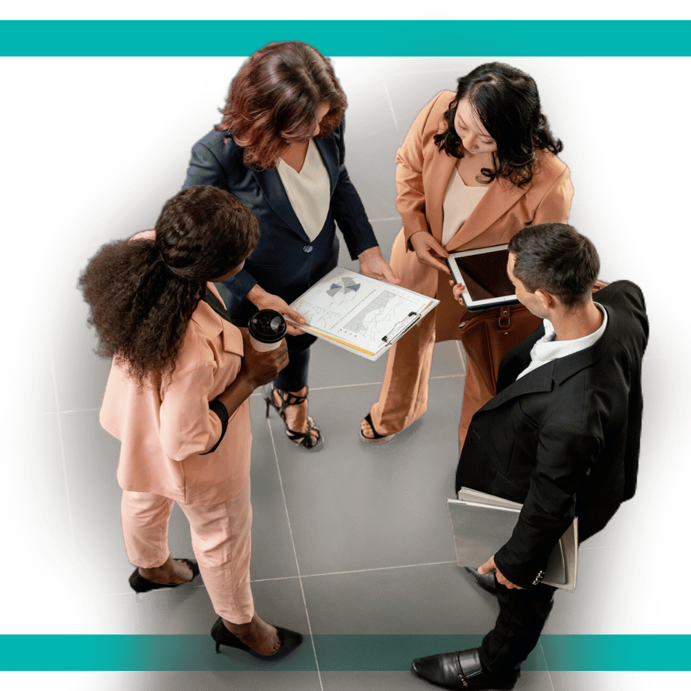 A group of four diverse business professionals standing in a collaborative discussion, reviewing a clipboard with charts and graphs, and using digital devices.