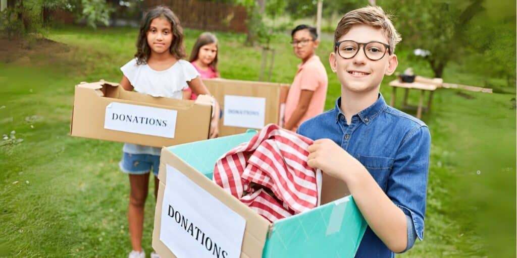 Children holding donation boxes filled with clothes and items in a grassy outdoor setting.